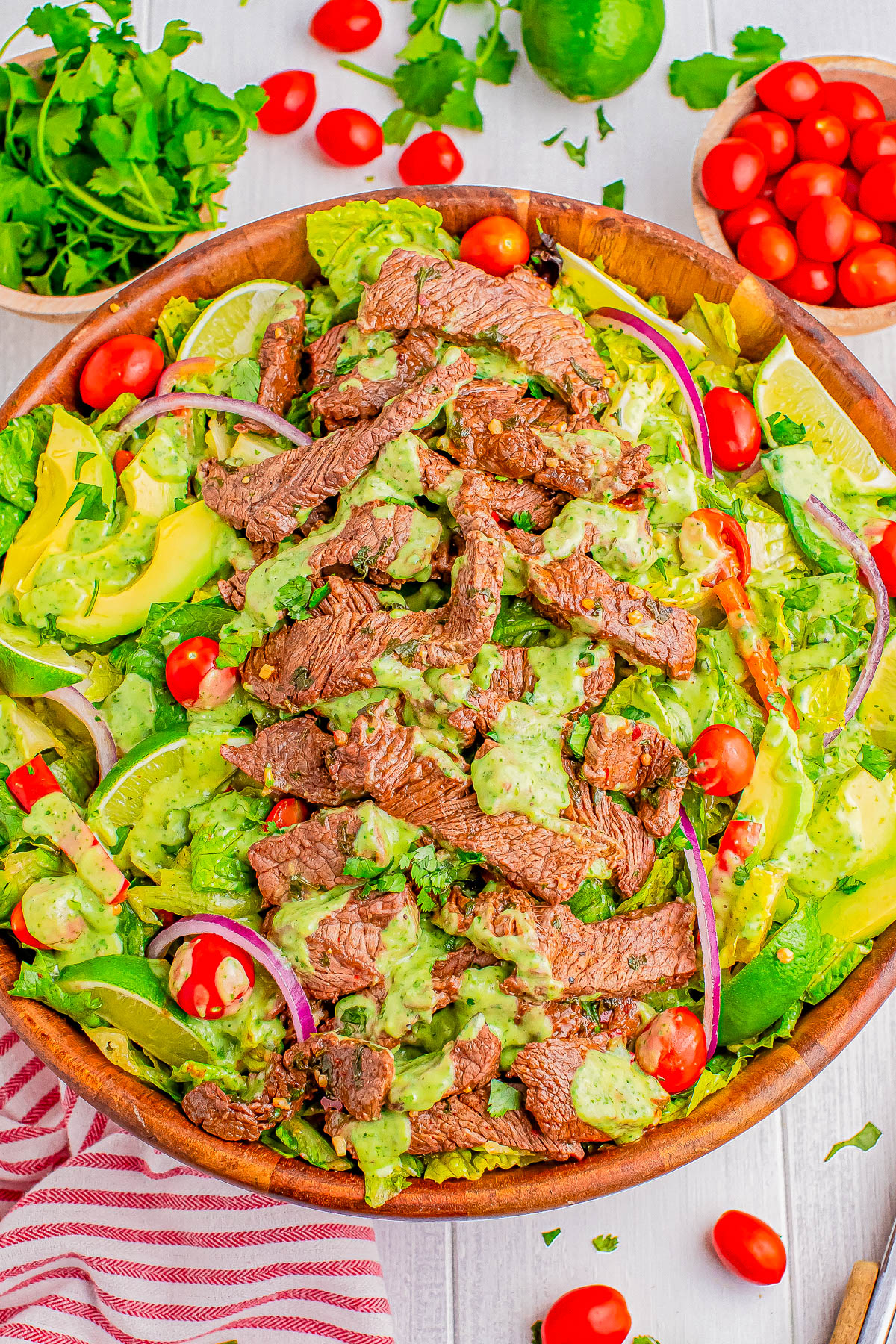 A wooden bowl filled with a salad featuring sliced steak, cherry tomatoes, avocado, red onion, and a green dressing. Other ingredients like parsley and lime are visible in the background.