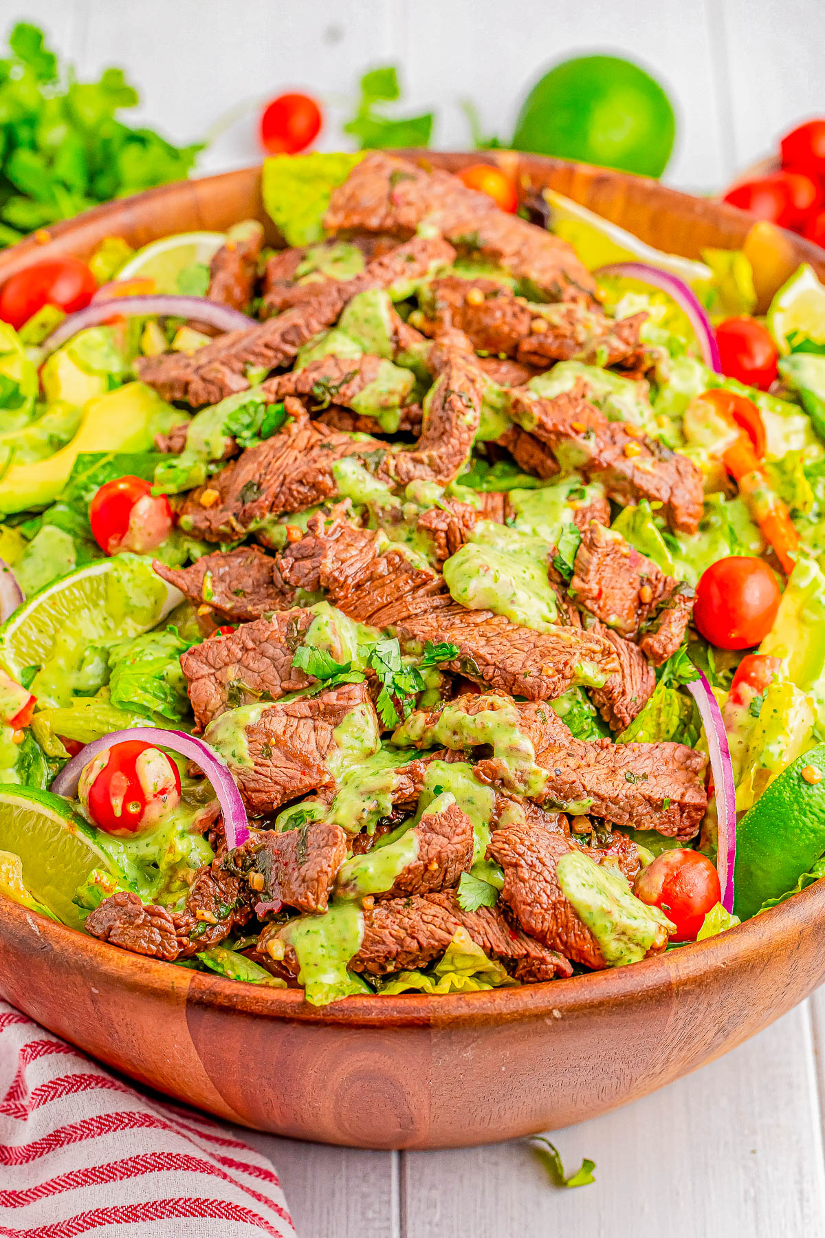 A wooden bowl containing a salad with steak strips, avocado slices, cherry tomatoes, red onion slices, and dressed with a green sauce.