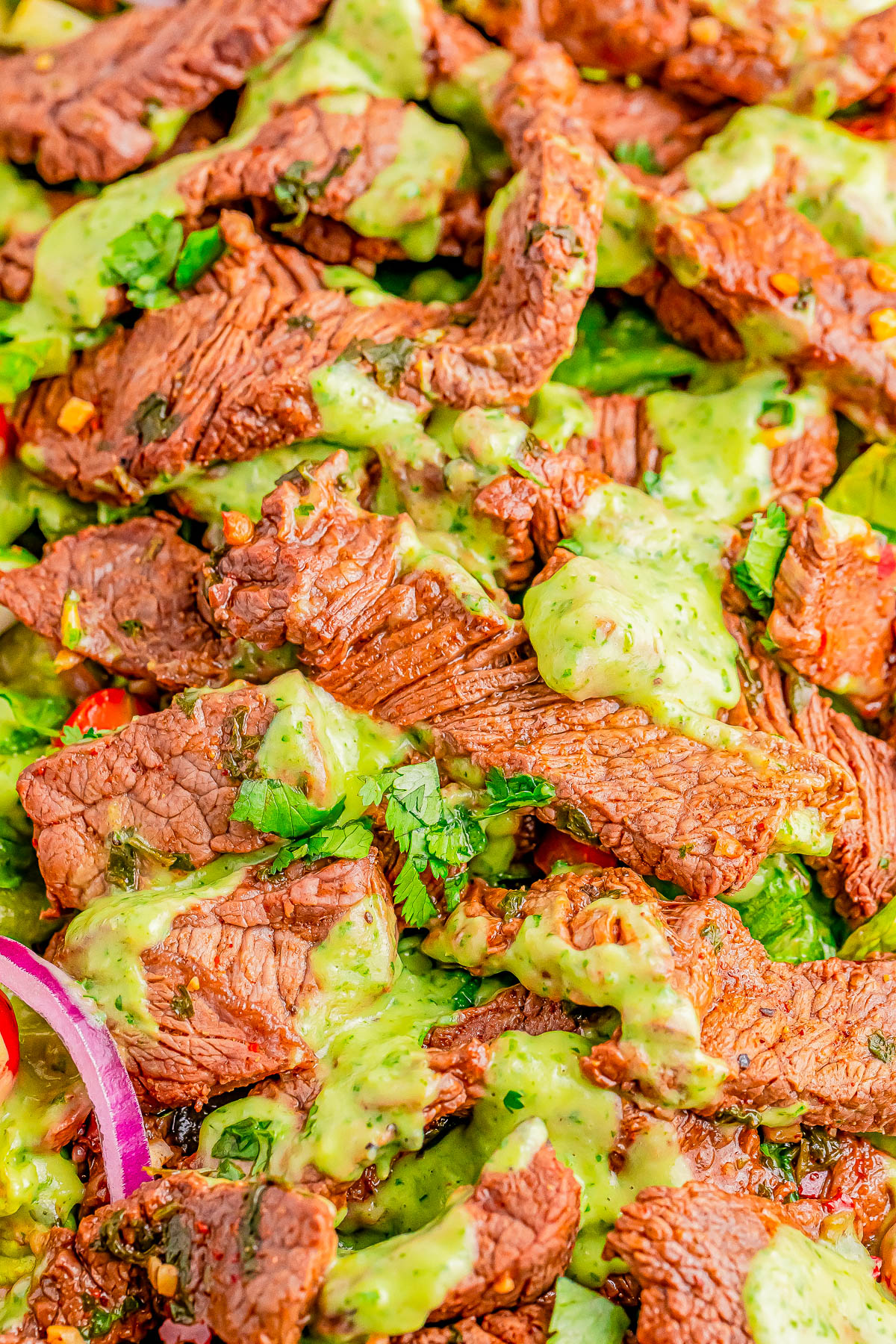 Close-up of sliced grilled beef topped with green sauce on a bed of fresh vegetables and herbs, including onions, cilantro, and cherry tomatoes.