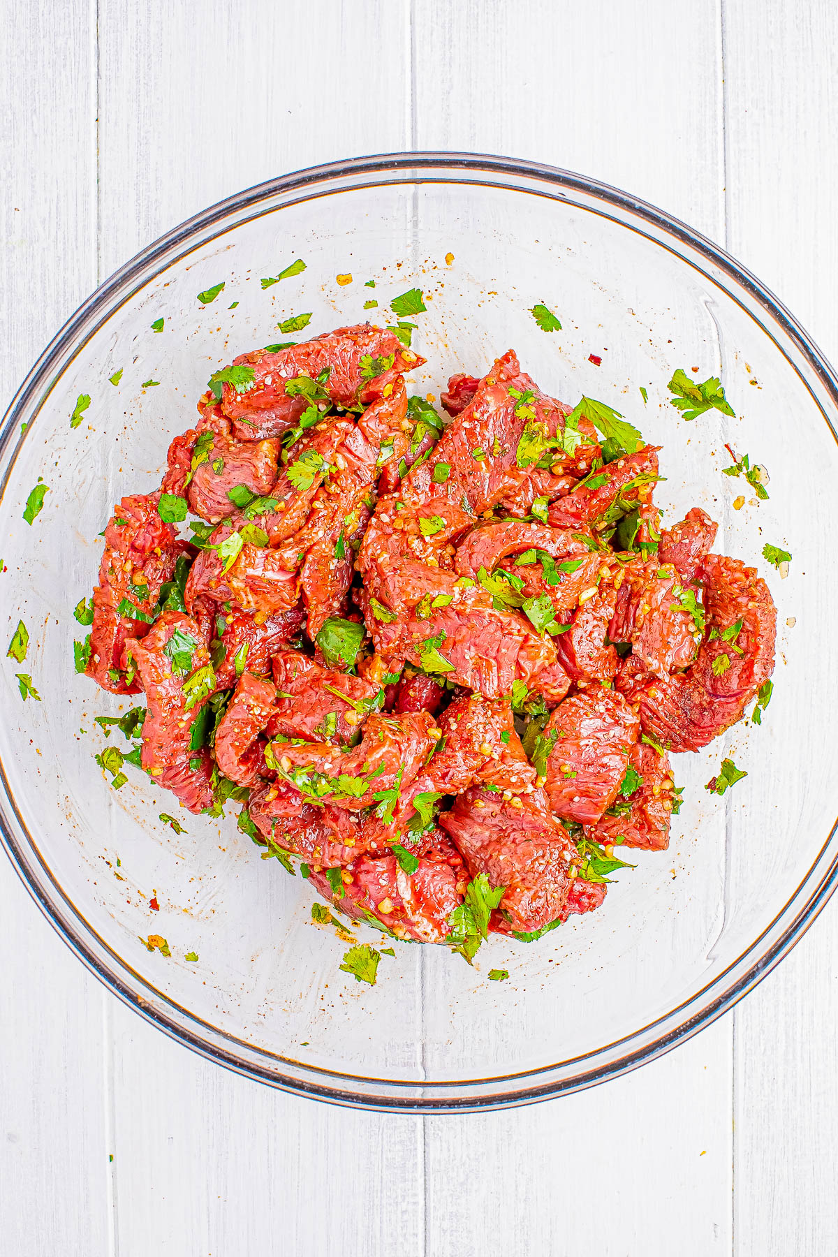 A glass bowl filled with marinated chunks of raw beef, garnished with chopped green herbs on a white wooden surface.