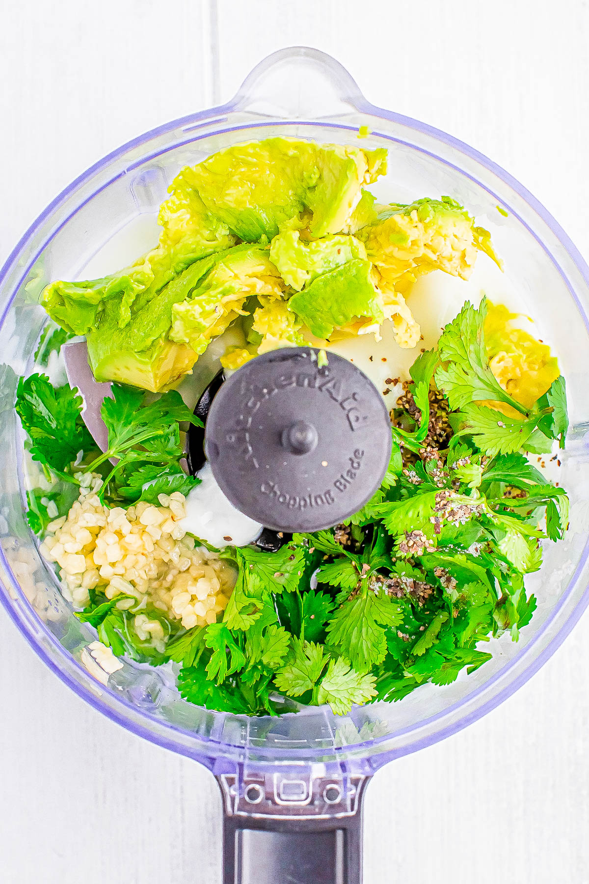 A food processor bowl filled with chopped avocado, cilantro, garlic, and seasonings, ready to be blended.