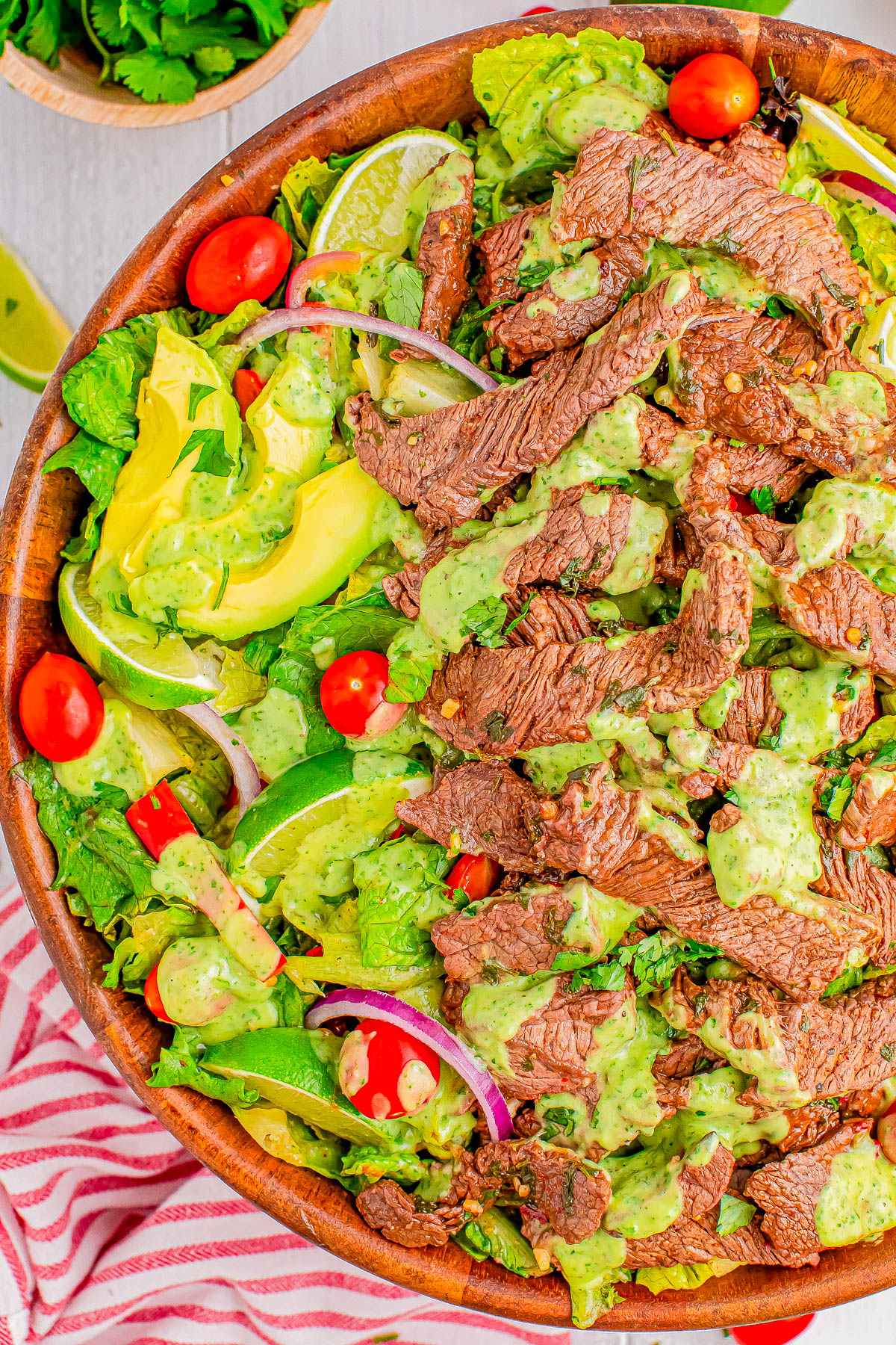 A wooden bowl filled with a mixed green salad topped with grilled steak, avocado, cherry tomatoes, red onion, lime wedges, and a creamy green dressing. A striped cloth is partially visible under the bowl.