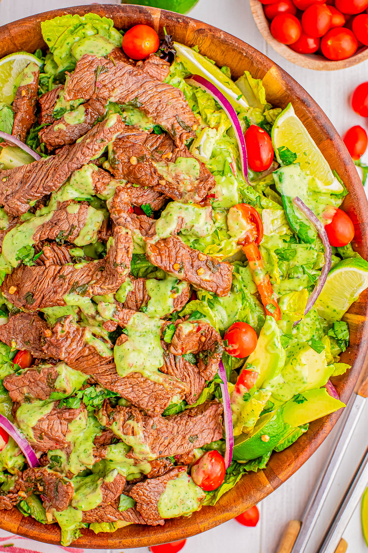 A wooden bowl filled with a steak salad featuring slices of steak, chopped lettuce, cherry tomatoes, red onion slices, avocado, and a drizzle of green dressing. Lime wedges garnished on the side.