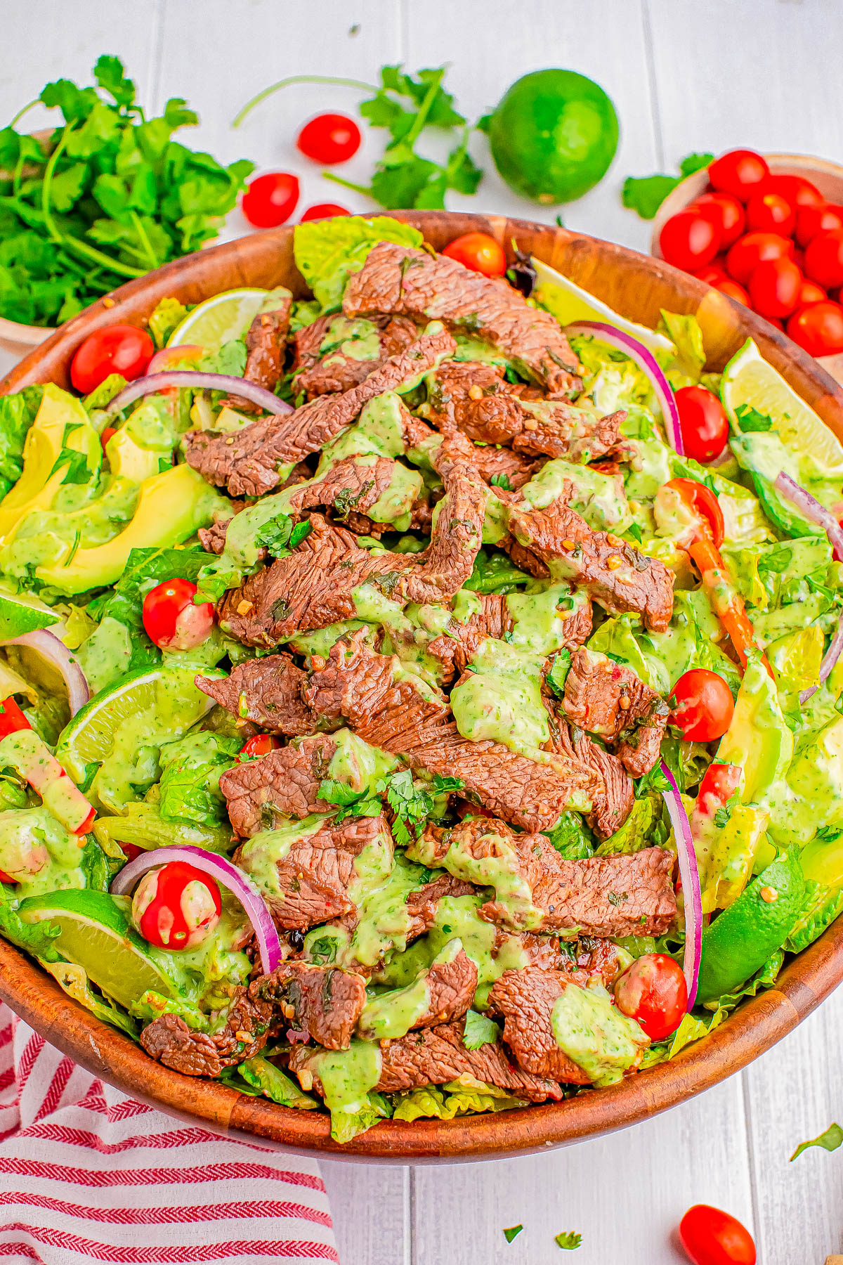 A wooden bowl filled with a salad of leafy greens, cucumbers, red onion slices, cherry tomatoes, avocado slices, topped with grilled steak strips, and drizzled with a green dressing.