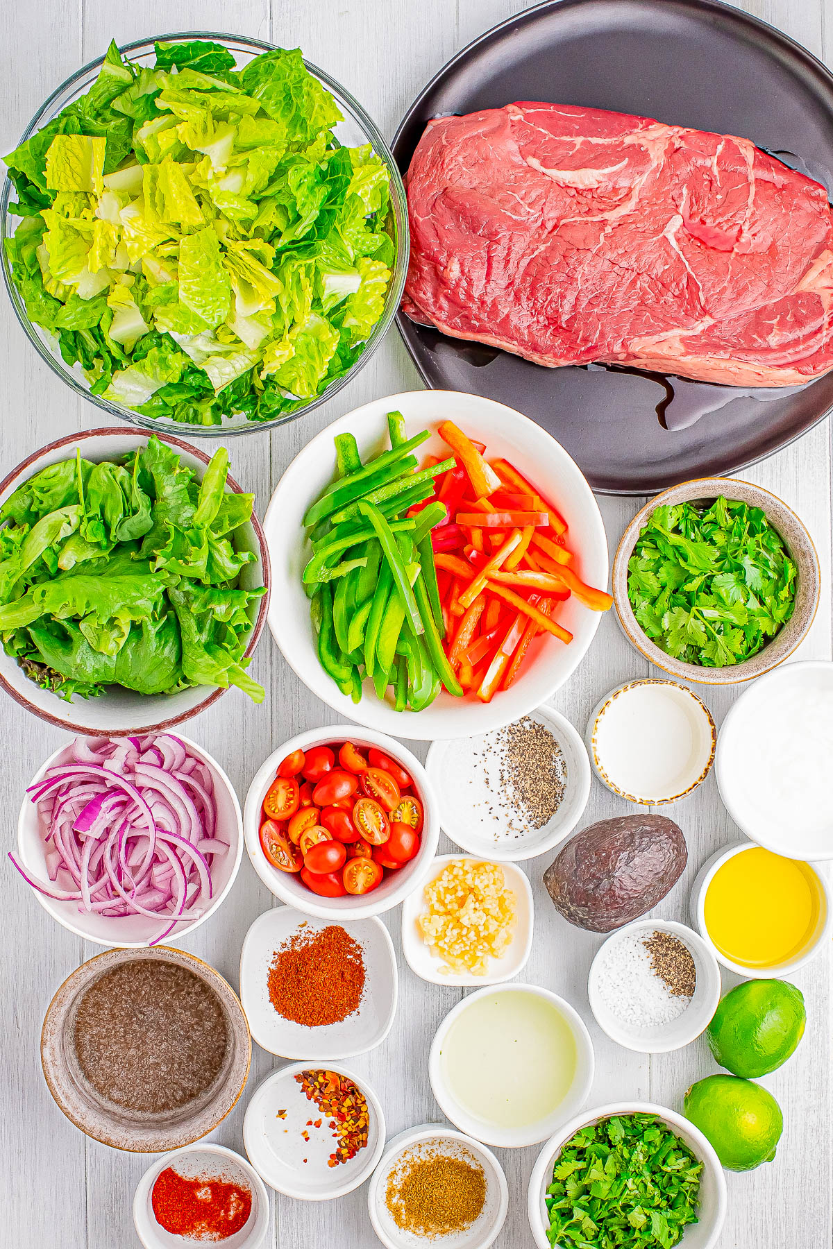 A display of various ingredients including a raw steak, chopped vegetables, sliced onions, herbs, spices, avocado, lime, and small bowls containing condiments and seasonings, arranged on a white surface.