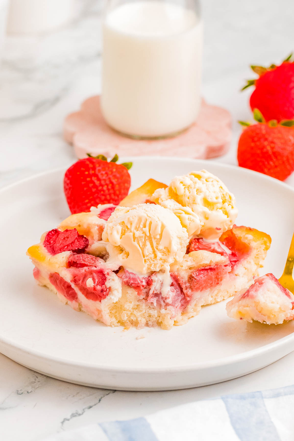 A slice of strawberry cobbler topped with vanilla ice cream on a white plate, accompanied by fresh strawberries and a glass of milk in the background.