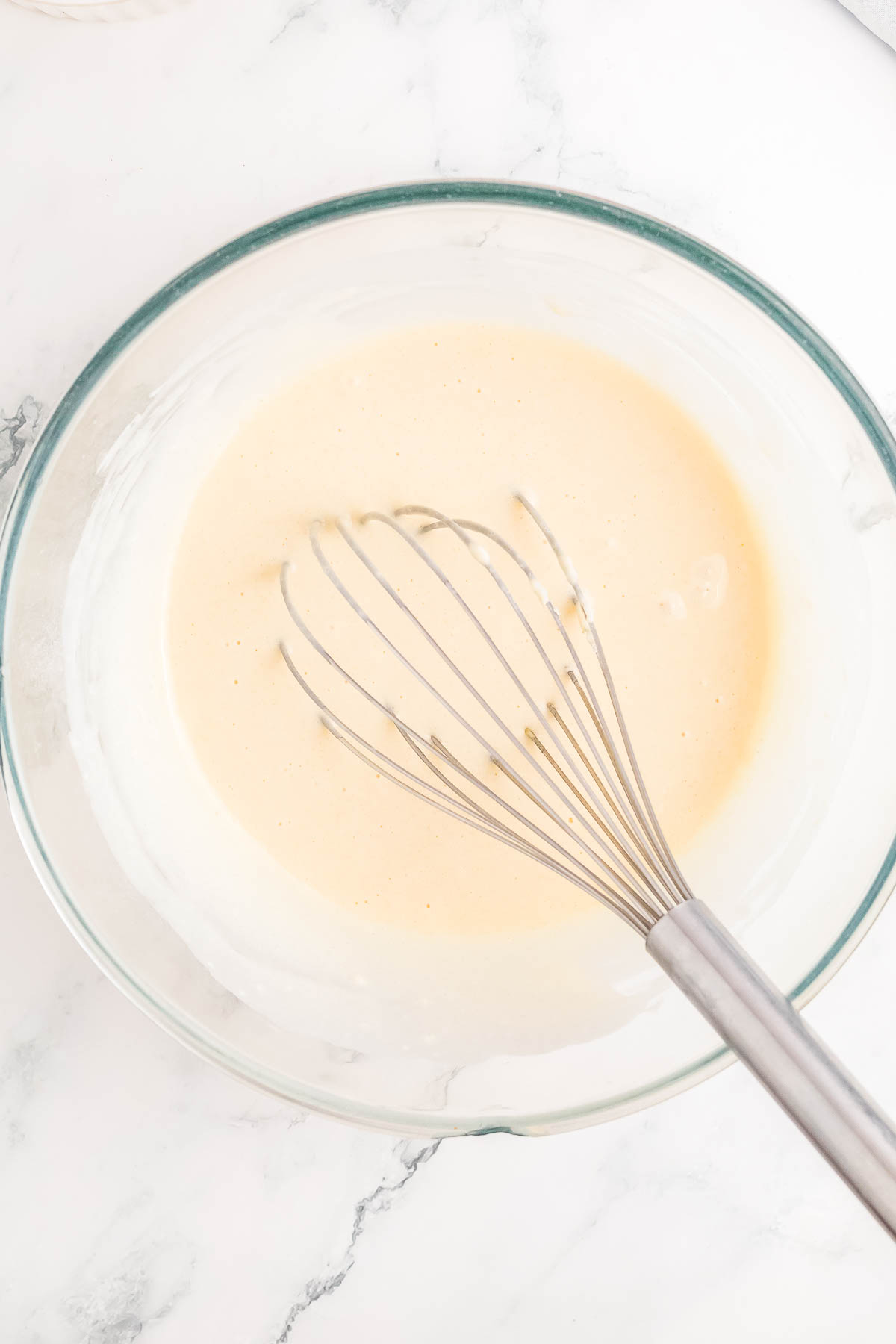 A clear glass bowl containing a light beige batter with a metal whisk, sitting on a marble surface.
