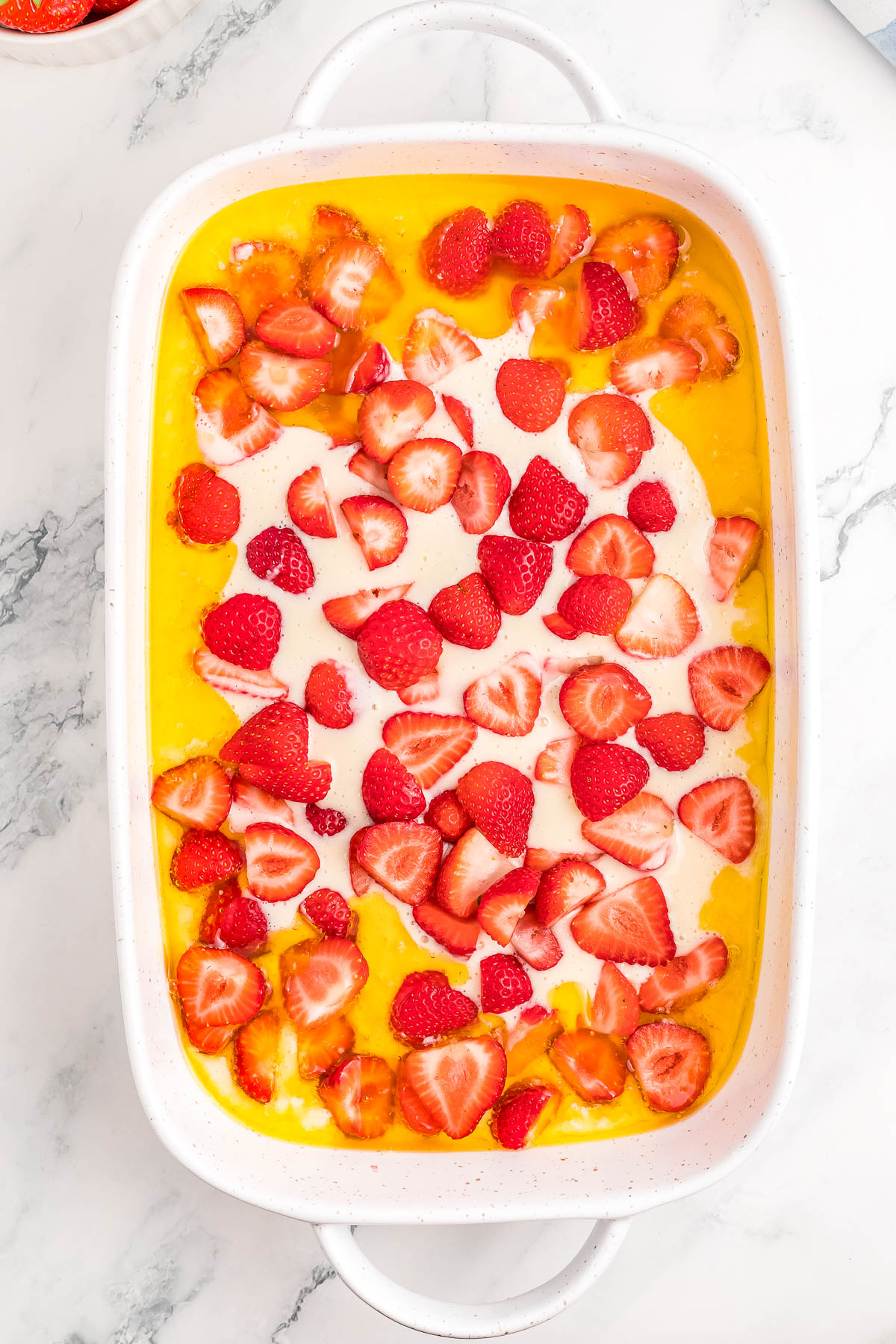 A dish of sliced strawberries on a bed of creamy custard in a white baking dish, viewed from above on a marble surface.