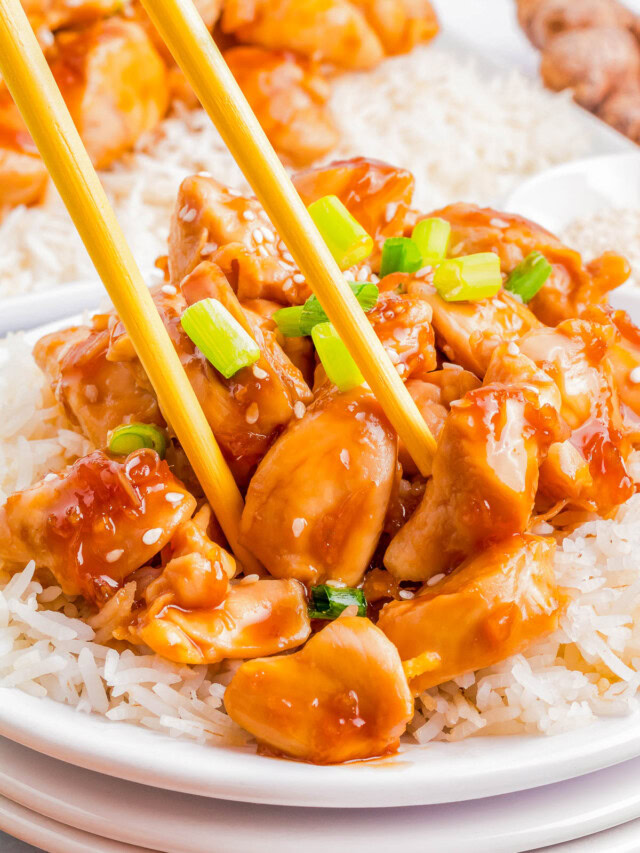 A close-up of a plate of rice topped with saucy chunks of teriyaki chicken, garnished with green onions, with chopsticks picking up a piece of chicken.