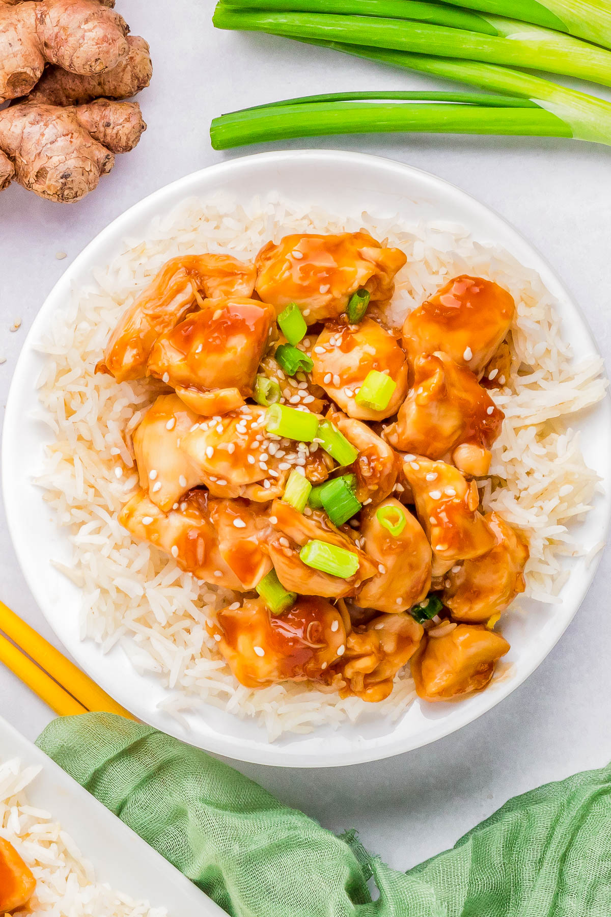 A bowl of white rice topped with pieces of orange-glazed chicken, garnished with sesame seeds and chopped green onions, with ginger root and green onions in the background.
