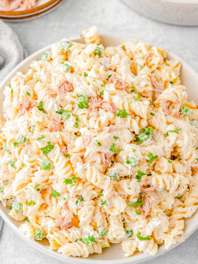 A bowl of creamy tuna pasta salad with rotini pasta, peas, and fresh parsley, placed on a light-colored surface.