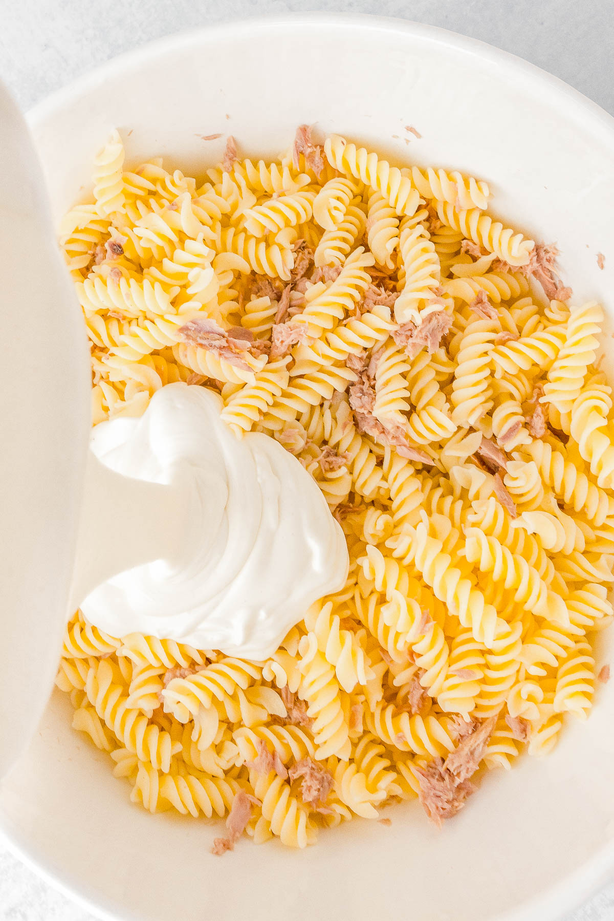 A bowl of rotini pasta mixed with shredded tuna, with a dollop of white creamy sauce being added on top.