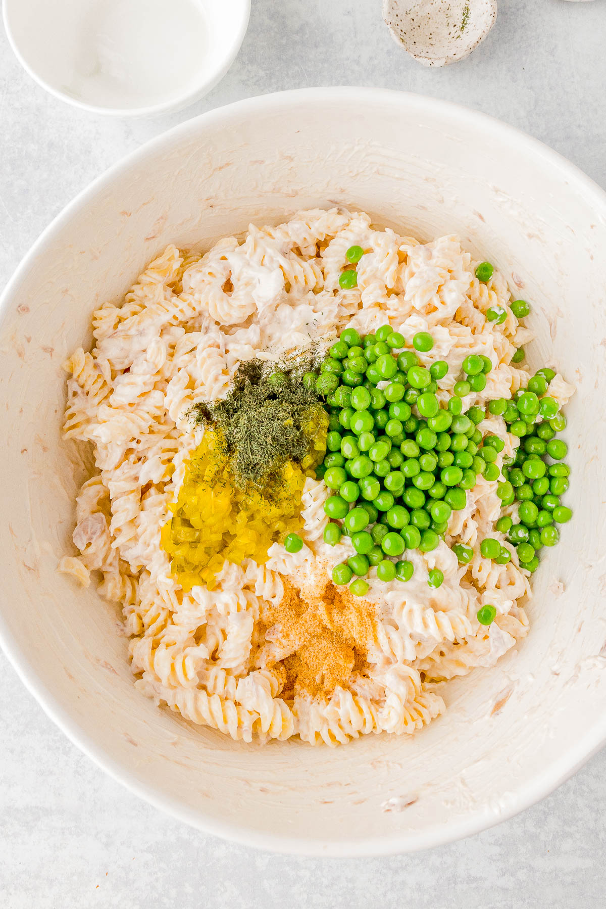 A bowl of cooked spiral pasta mixed with peas, seasonings, and a creamy dressing, placed on a white surface.