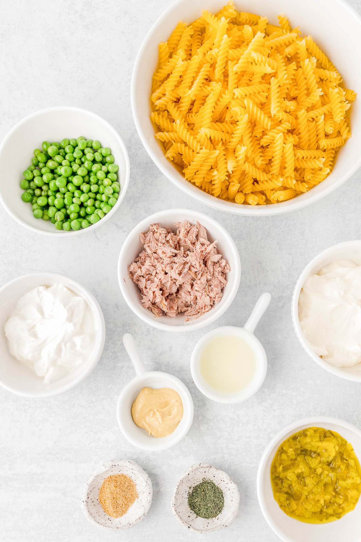 Ingredients for a pasta salad: uncooked pasta spirals, peas, shredded meat, mayonnaise, sour cream, mustard, lemon juice, and spices neatly arranged in bowls on a white surface.