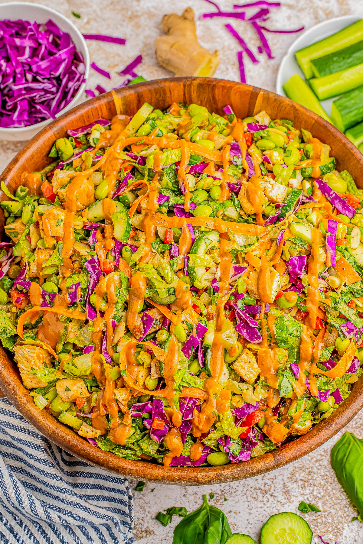 A large wooden bowl filled with a colorful salad of mixed greens, purple cabbage, edamame, and other vegetables, topped with an orange dressing. Plates of additional ingredients are in the background.