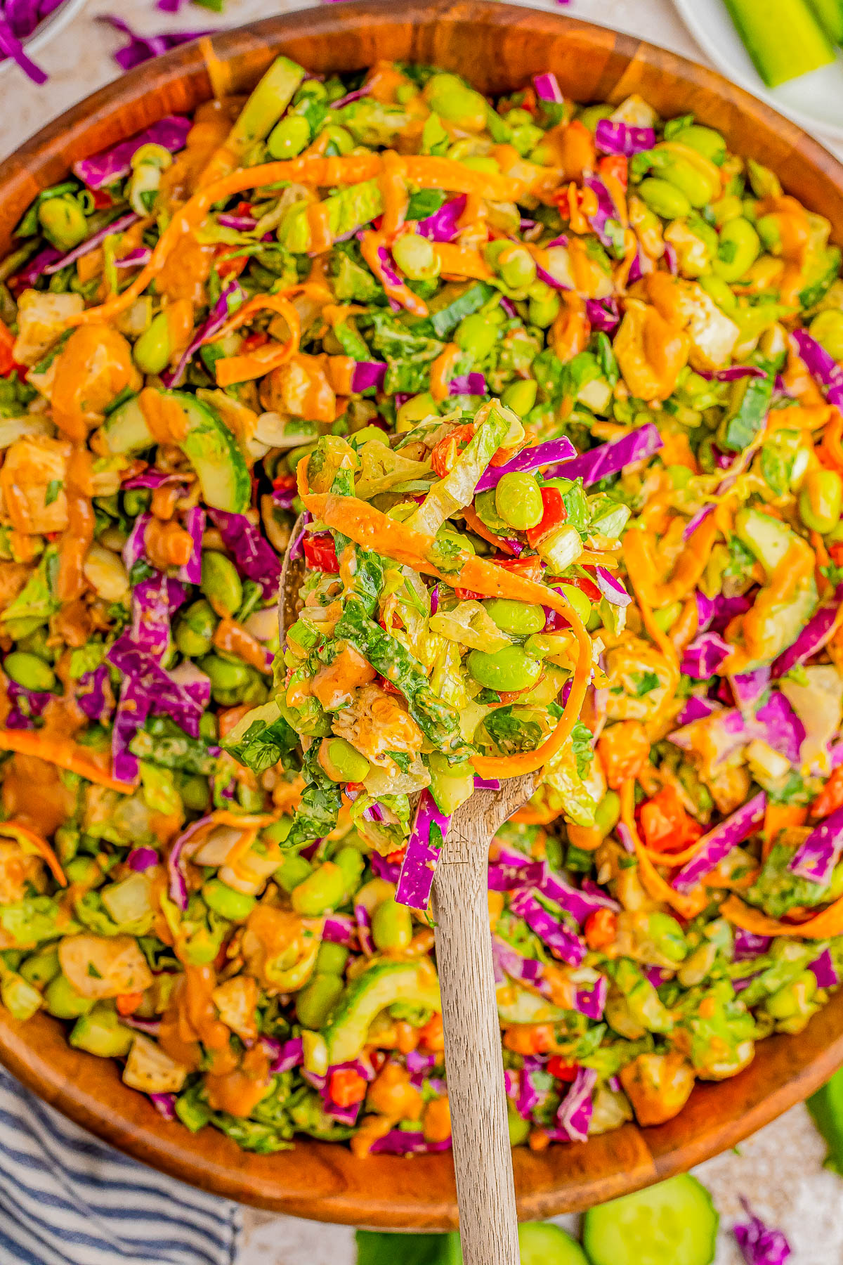 A wooden spoon holds a portion of colorful salad with ingredients like lettuce, cucumber, edamame, carrots, and purple cabbage, dressed in an orange-colored sauce, from a large wooden bowl.