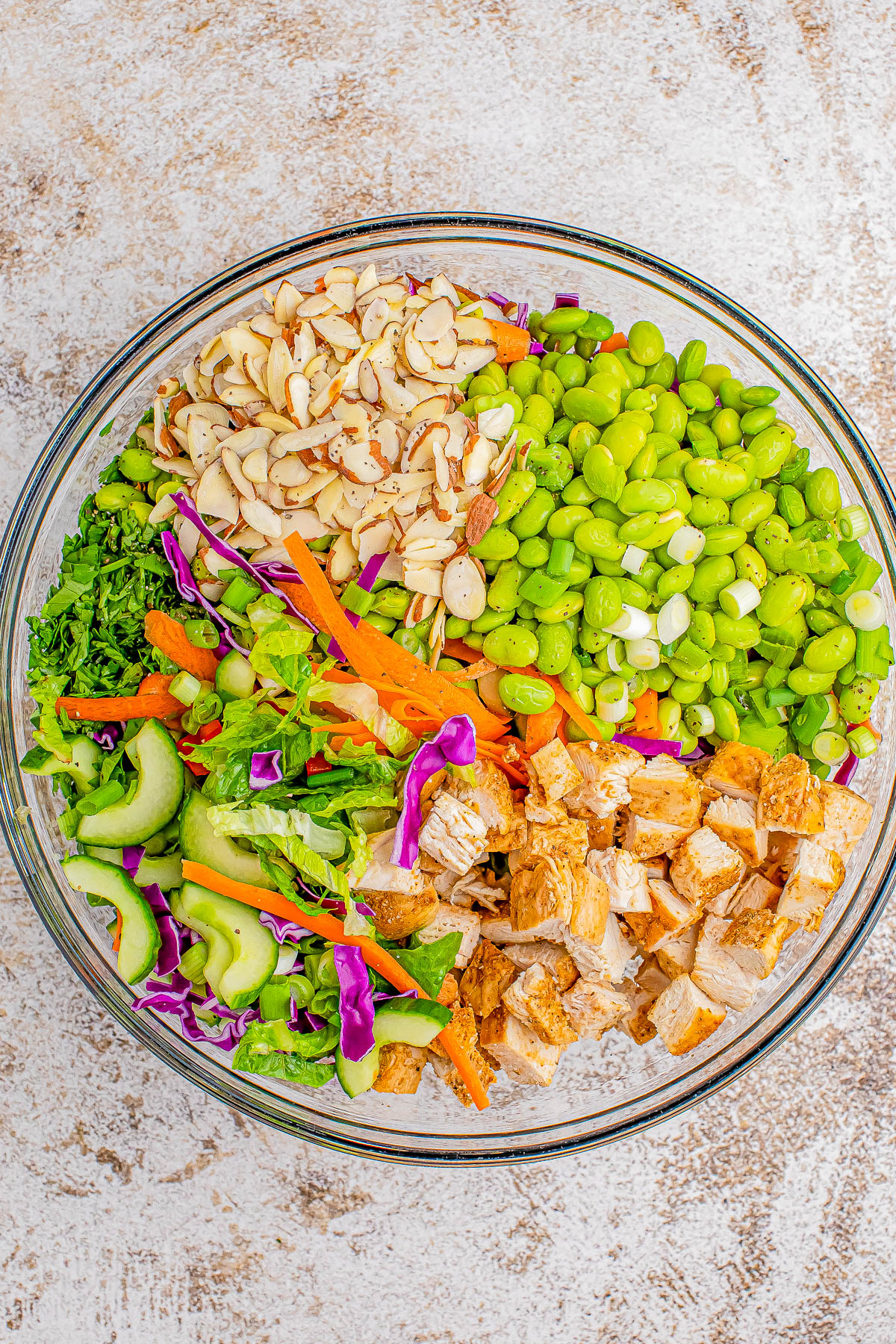 A glass bowl filled with a colorful salad consisting of sliced almonds, edamame, chopped cooked chicken, cucumber, red cabbage, carrots, and mixed greens.