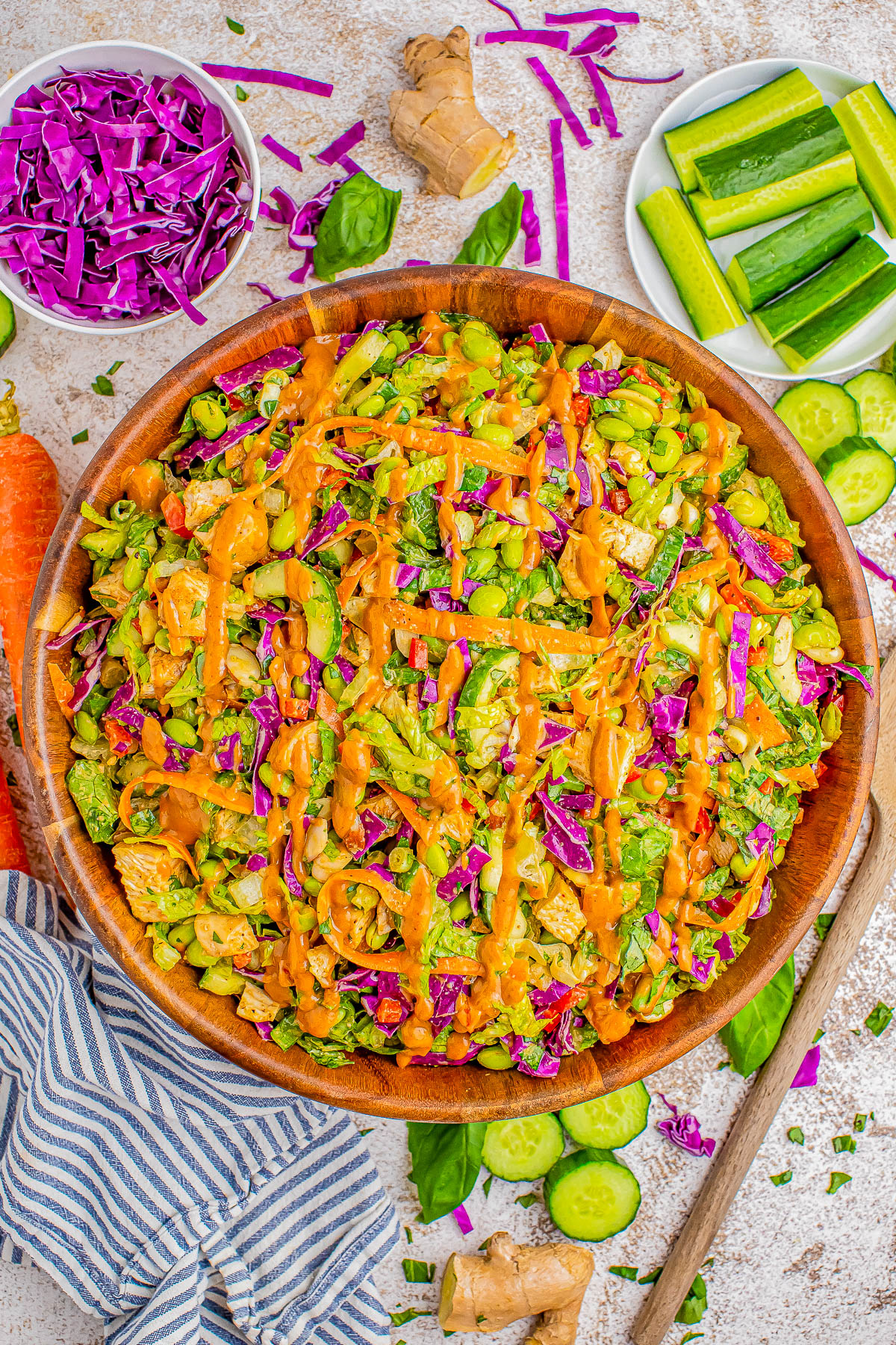 A colorful salad in a wooden bowl, featuring lettuce, purple cabbage, cucumber, carrots, edamame, and orange dressing. Plates with cucumber sticks and shredded purple cabbage are on the side. A striped cloth is nearby.