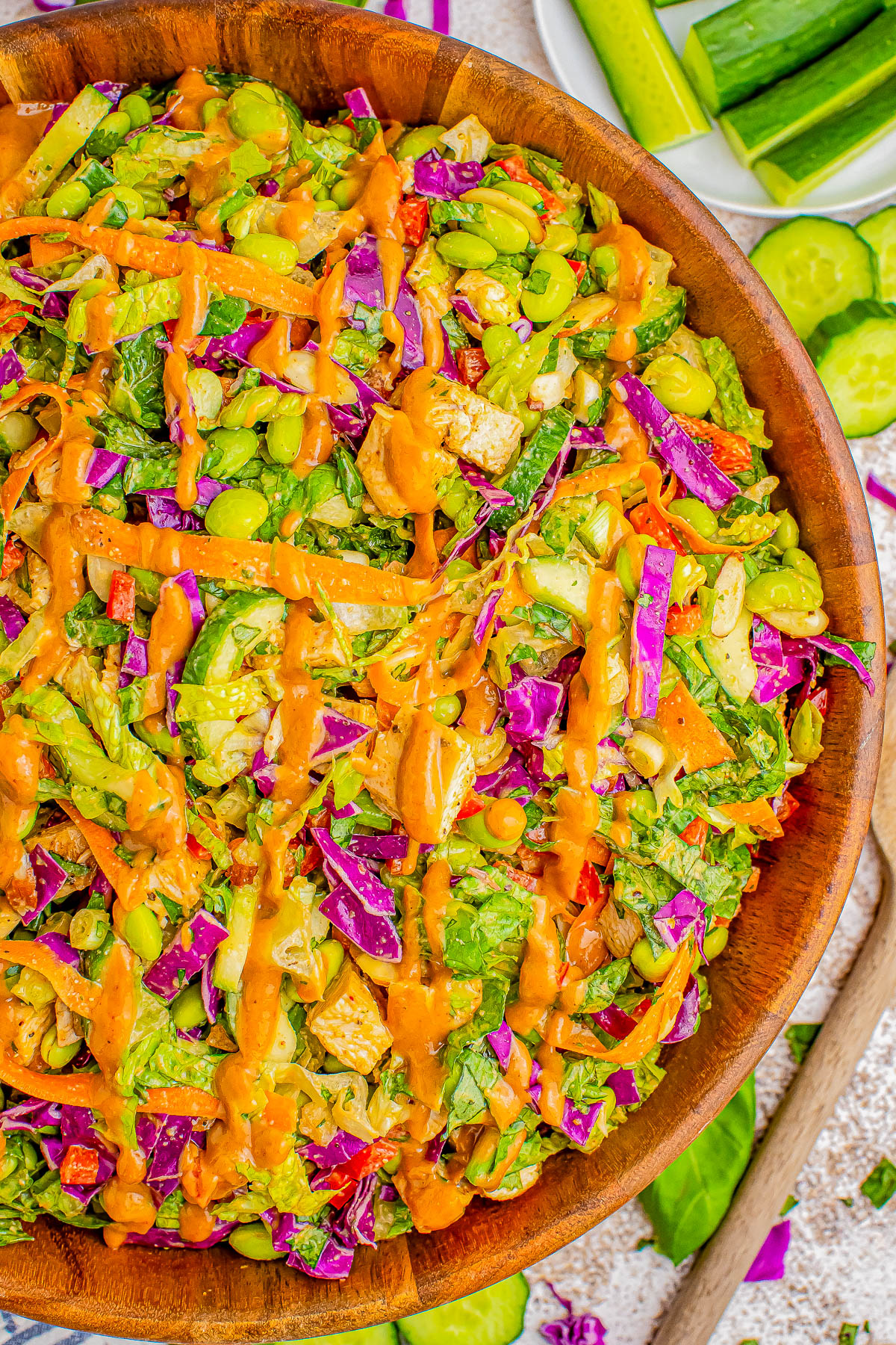 A wooden bowl filled with a vibrant mixed salad featuring chopped vegetables and a drizzle of orange dressing. Slices of cucumber and chopped vegetables are visible in the background.