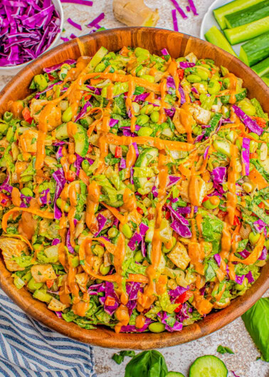 A wooden bowl filled with a colorful vegetable salad topped with a drizzle of orange dressing. Sliced cucumbers, chopped red cabbage, greens, and ginger surround the bowl.