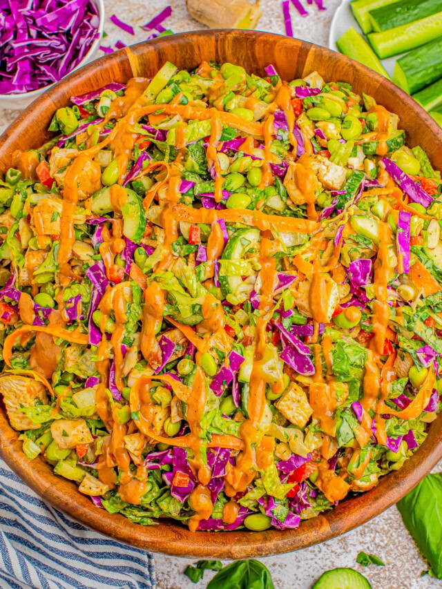 A wooden bowl filled with a colorful vegetable salad topped with a drizzle of orange dressing. Sliced cucumbers, chopped red cabbage, greens, and ginger surround the bowl.