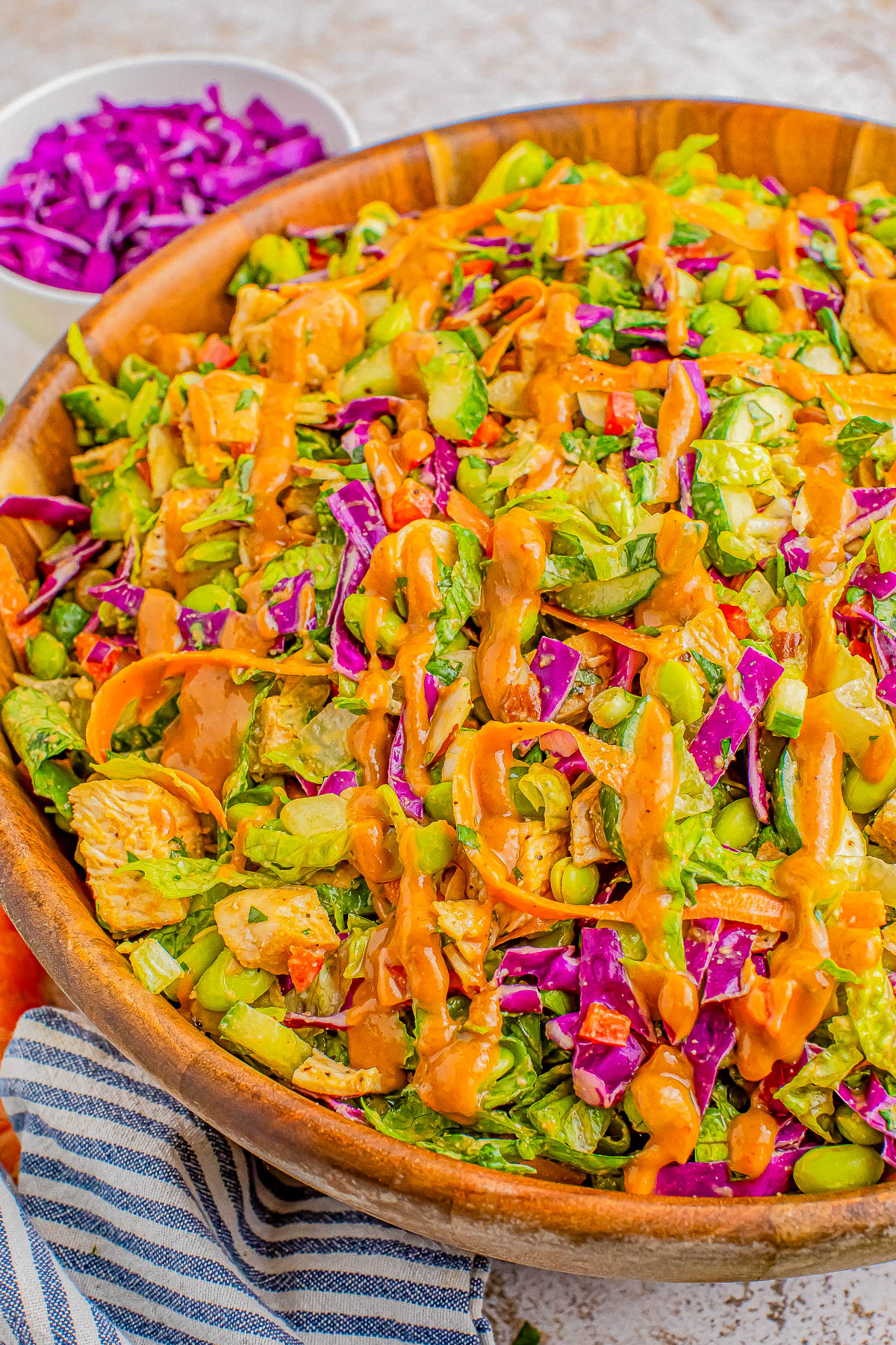 A close-up of a colorful salad in a wooden bowl, topped with a drizzle of creamy dressing. The salad includes red cabbage, edamame, and greens. A small bowl of shredded red cabbage is in the background.