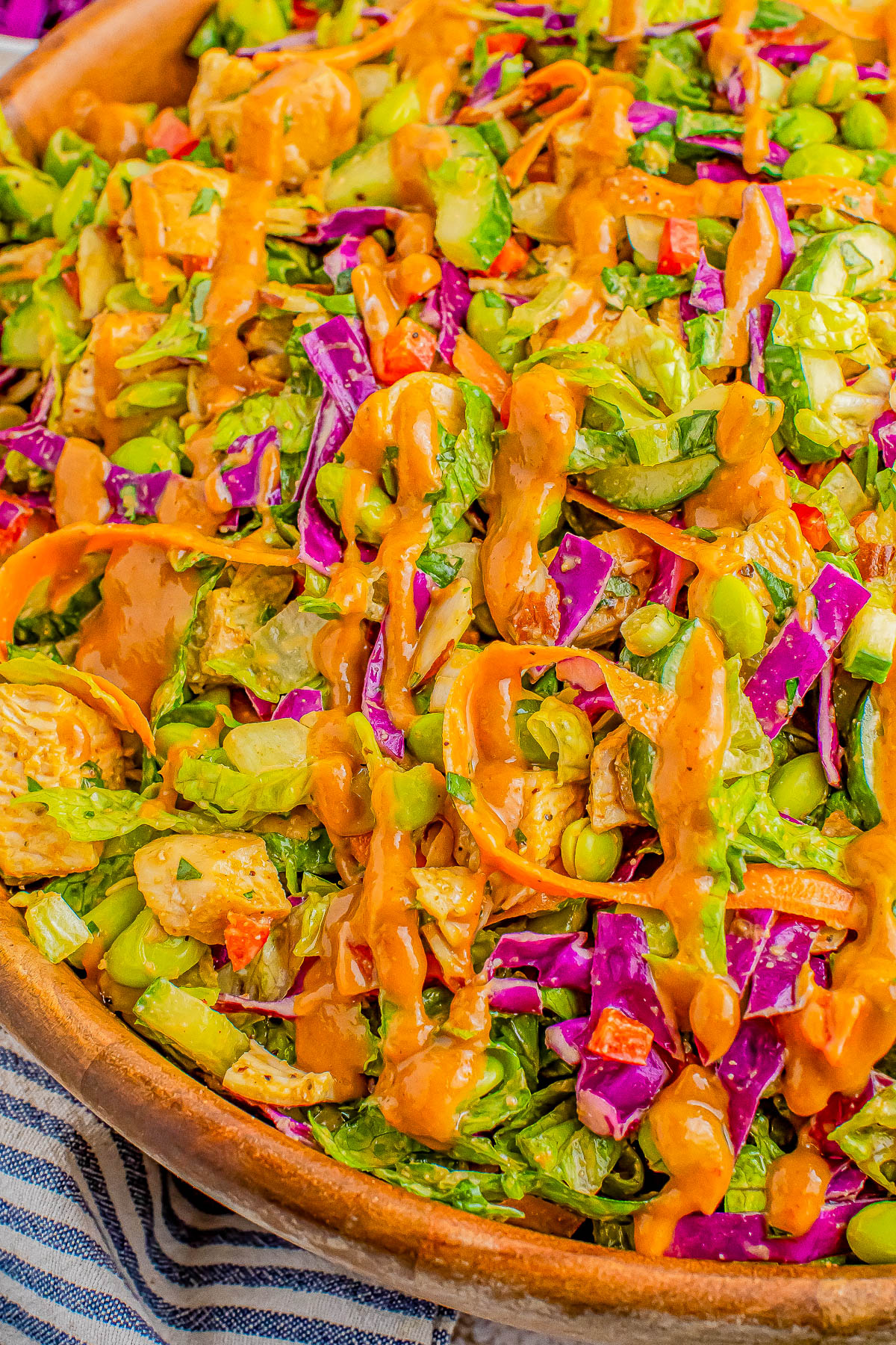 A close-up of a colorful salad with chopped vegetables and mixed greens, topped with a drizzle of dressing. The salad includes carrots, purple cabbage, edamame, and other assorted vegetables.