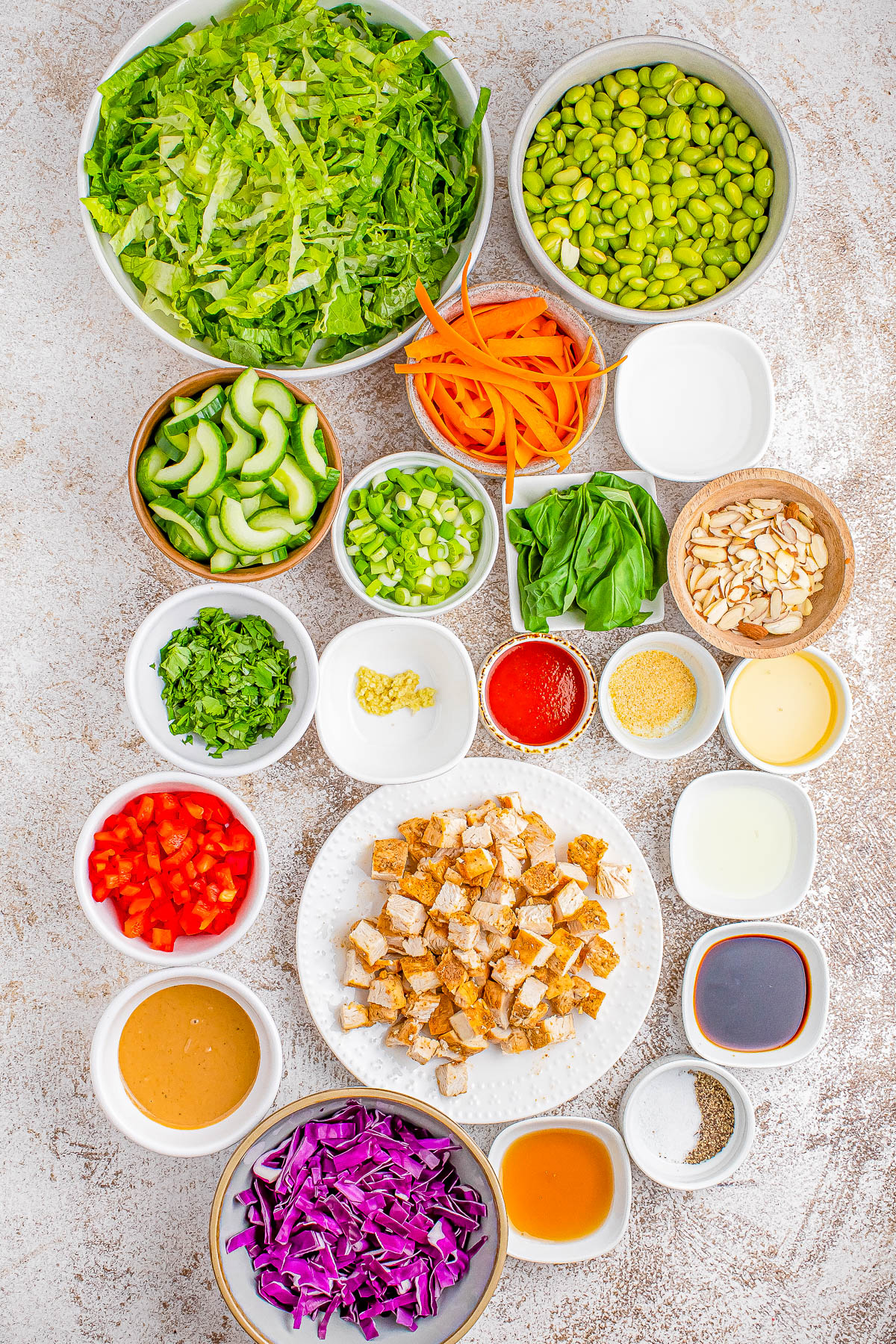 An assortment of vegetables, tofu, and condiments are arranged in small bowls. The ingredients include lettuce, edamame, carrots, cucumbers, bell peppers, purple cabbage, various sauces, and seasonings.