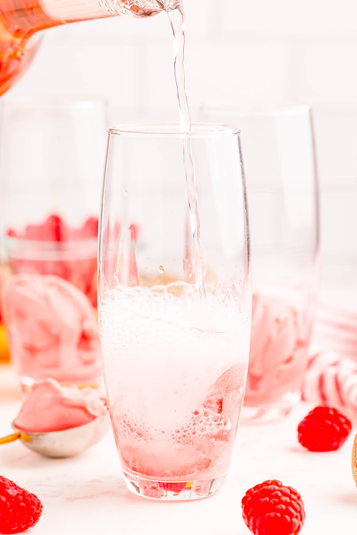 A close-up of a drink being poured into a glass containing a scoop of raspberry sorbet. Other similar glasses with sorbet and fresh raspberries are in the background.