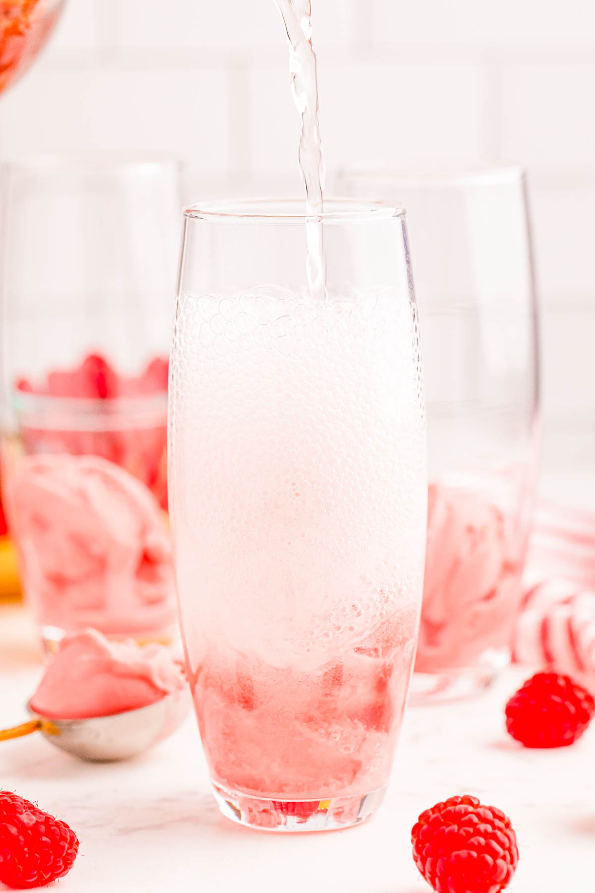 A clear glass being filled with a pink foamy liquid, likely raspberry-flavored, with fresh raspberries and scoops of pink ice cream visible at the bottom.