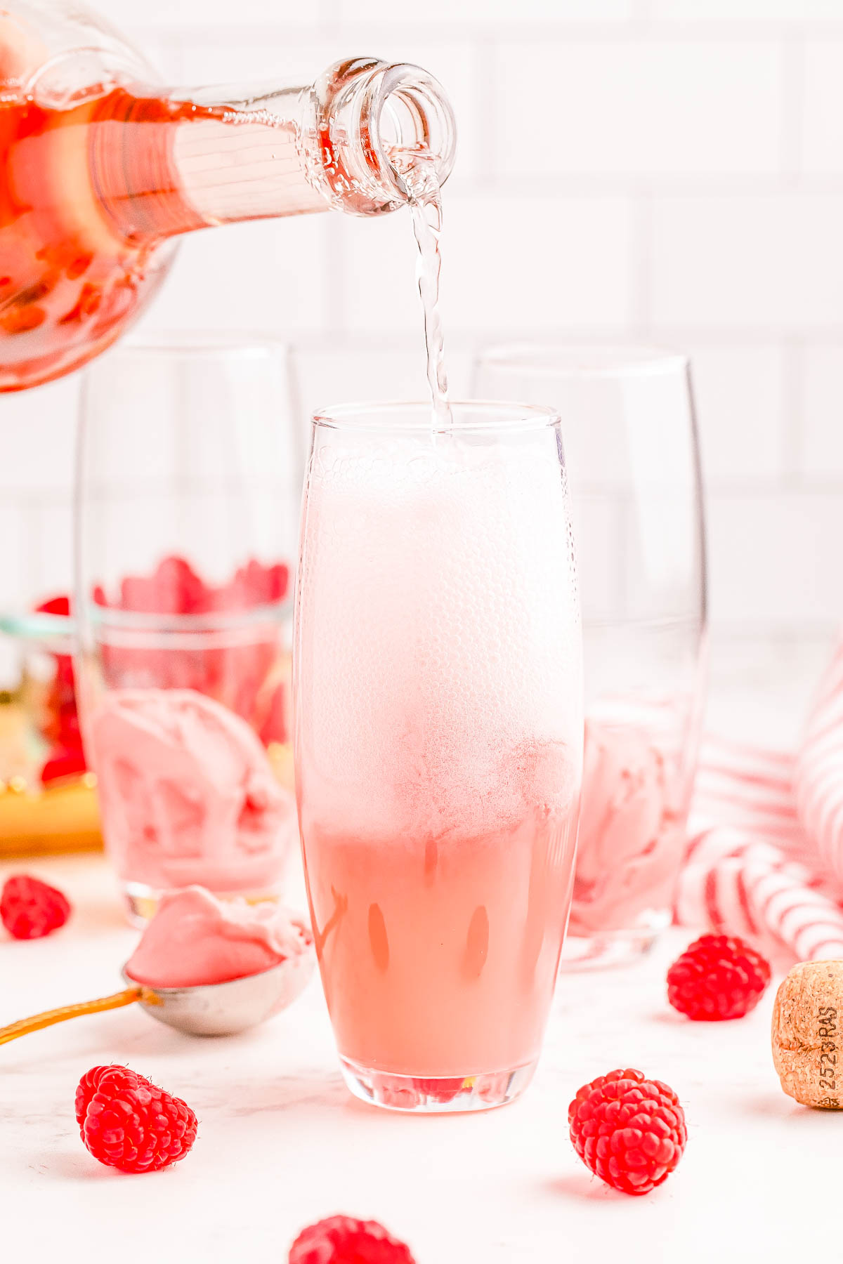 A bottle of rosé champagne is being poured into a tall glass, causing it to foam. Fresh raspberries are scattered on the table and a spoon with raspberry sorbet is nearby.