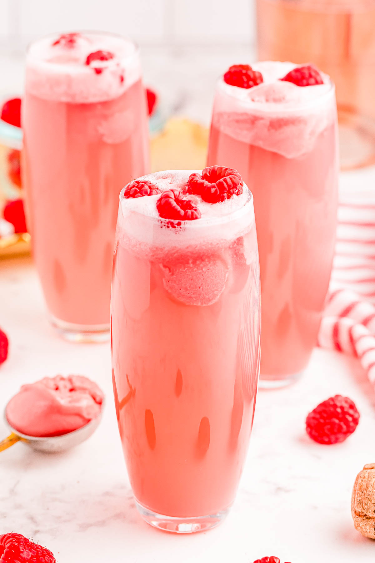 Three tall glasses of pink punch topped with raspberries and foam, surrounded by scattered raspberries and a spoonful of pink sorbet on a white counter.