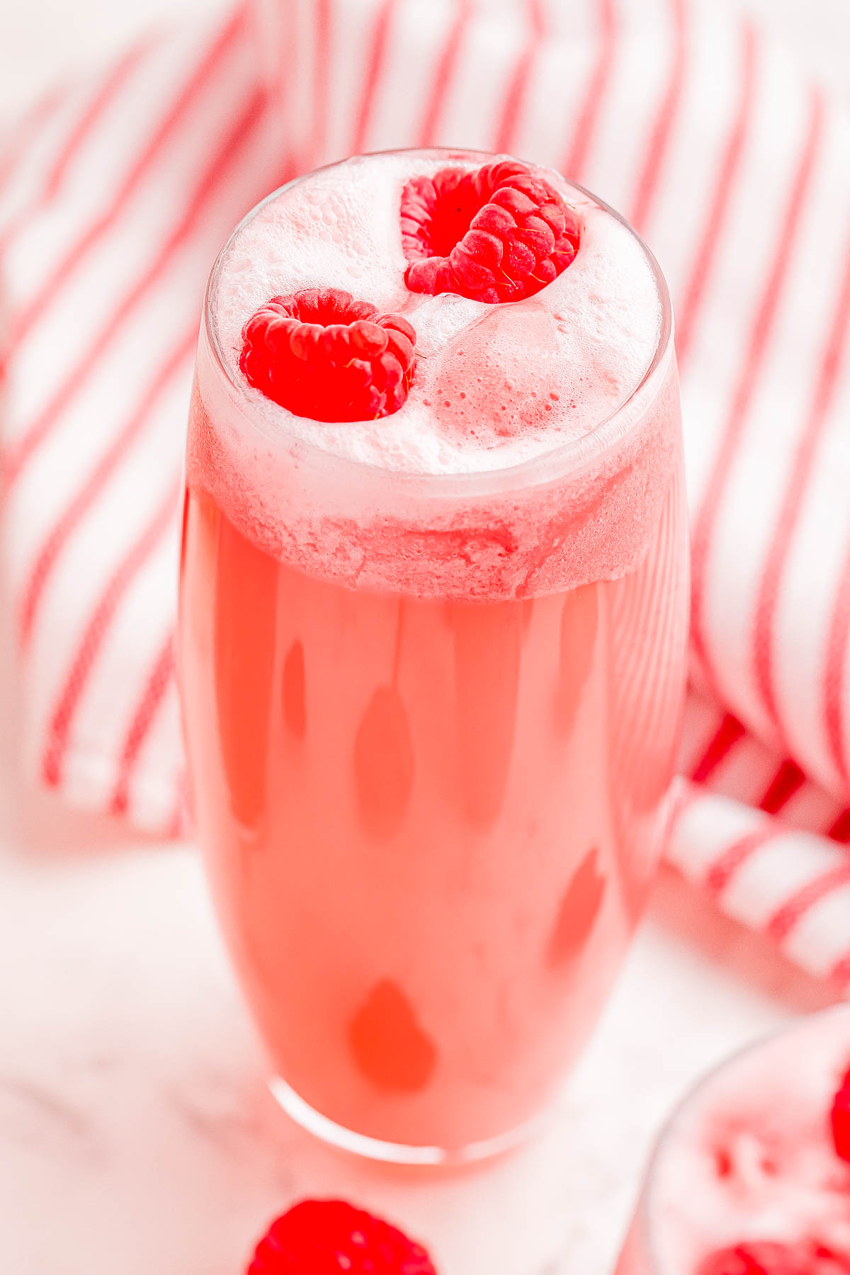 A tall glass filled with a frothy pink beverage, garnished with fresh raspberries. A red and white striped cloth is visible in the background.