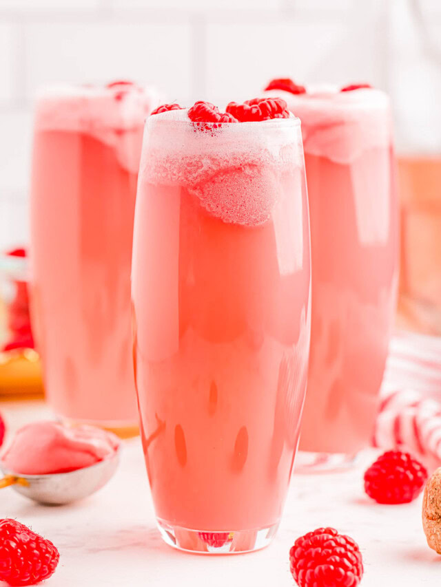 Three tall glasses filled with a frothy pink beverage, topped with raspberries, surrounded by scattered raspberries and a glimpse of a spoon with a pink substance.