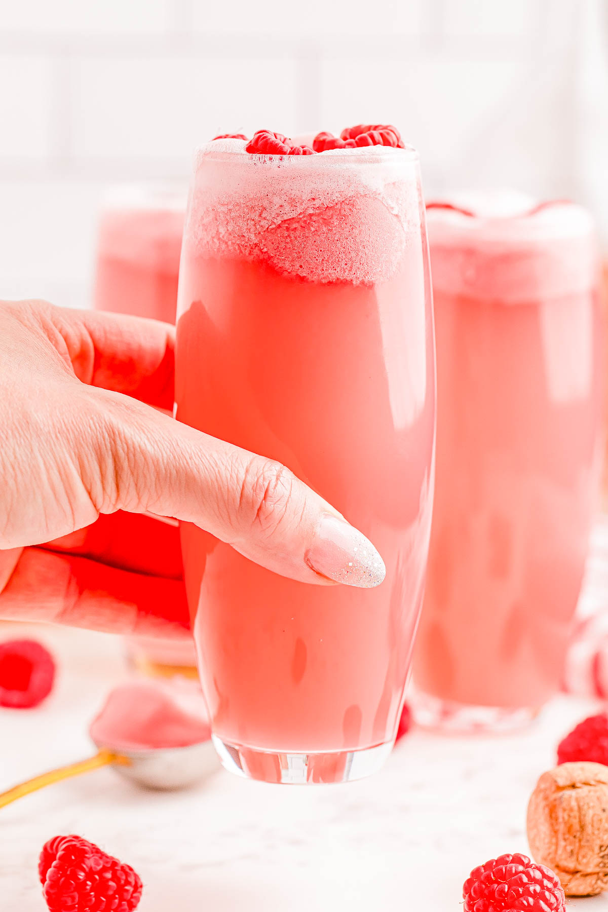 A hand holds a tall glass filled with pink beverage and garnished with raspberries. Several similar glasses and scattered raspberries are seen in the background.