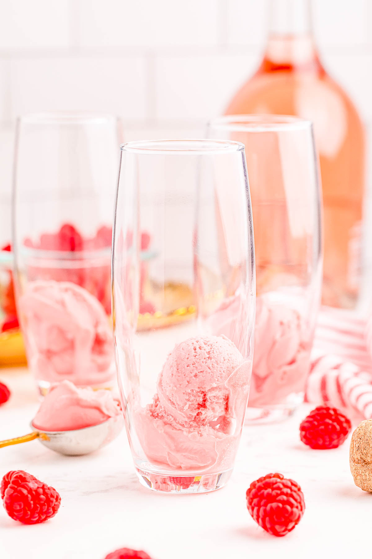 Three tall glasses each containing a scoop of pink ice cream, surrounded by fresh raspberries and an open bottle of rosé in the background.