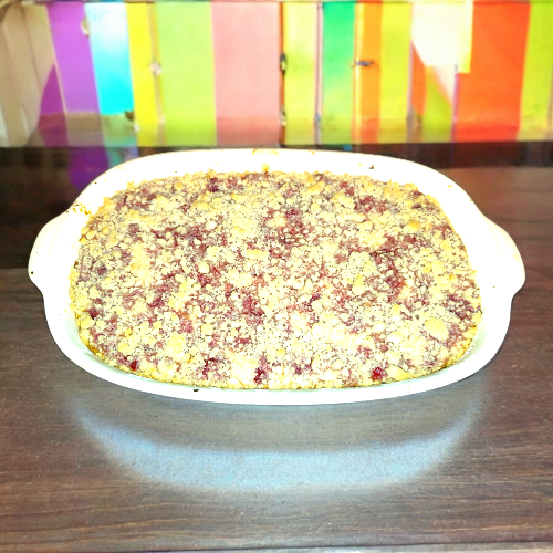 A white baking dish filled with a fruit crumble dessert, placed on a dark wooden surface with a colorful striped background.