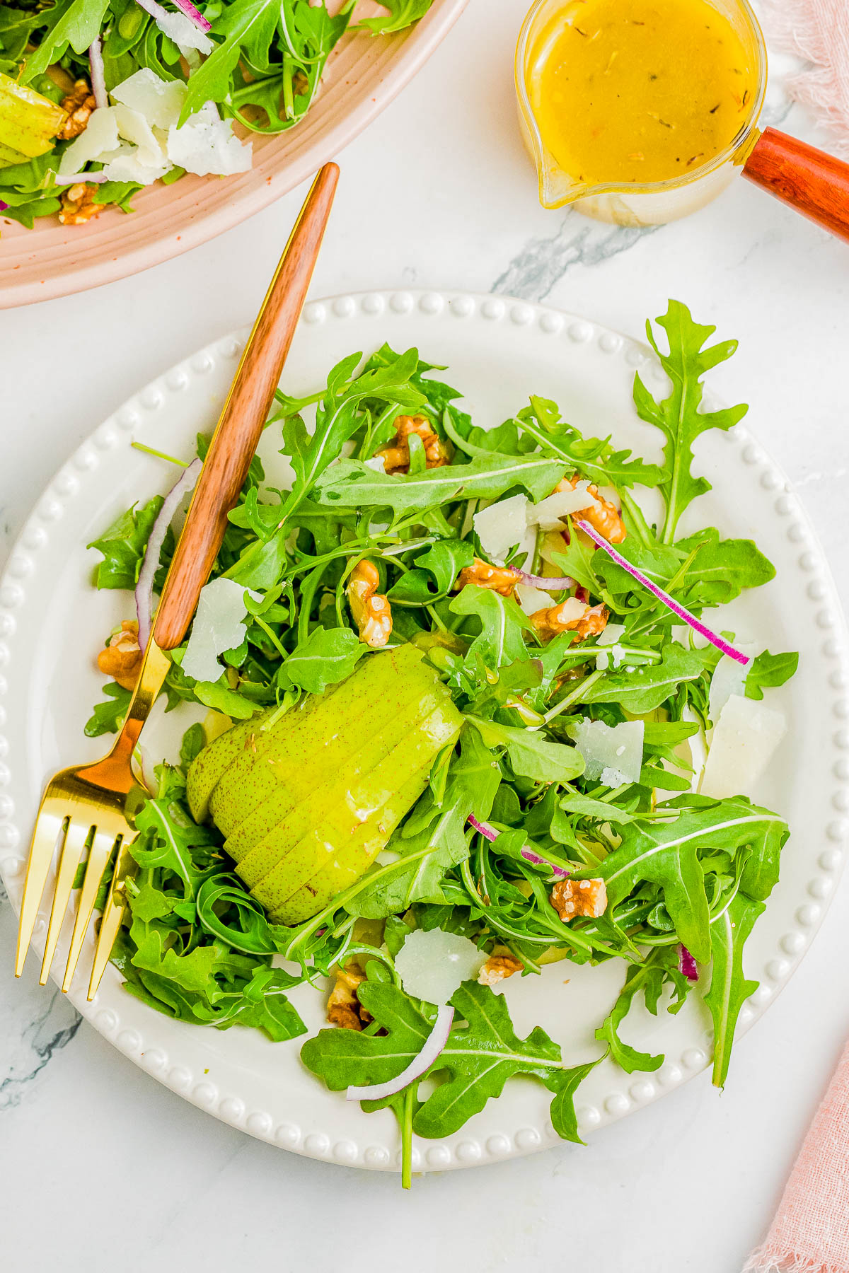 A fresh arugula salad on a white plate, topped with avocado slices, walnuts, shaved cheese, and red onion, served with a fork and knife. A small container of dressing is placed beside the plate.