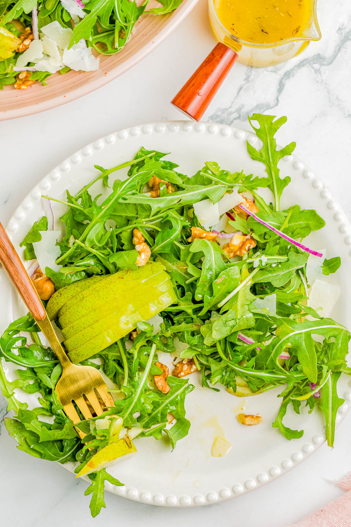 A plate of arugula salad with pear slices, walnuts, shaved cheese, and red onion, accompanied by a fork and a small container of dressing on the side.