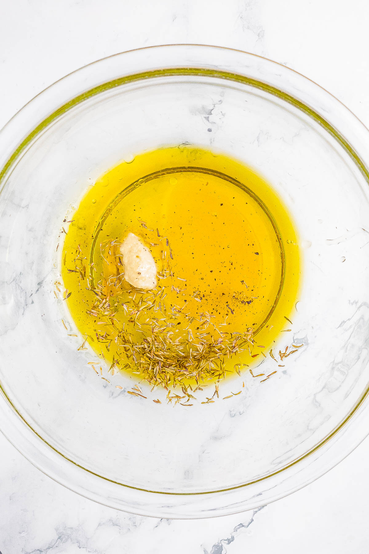 A clear glass bowl containing olive oil, a dollop of mustard, dried herbs, and black pepper on a marble surface.