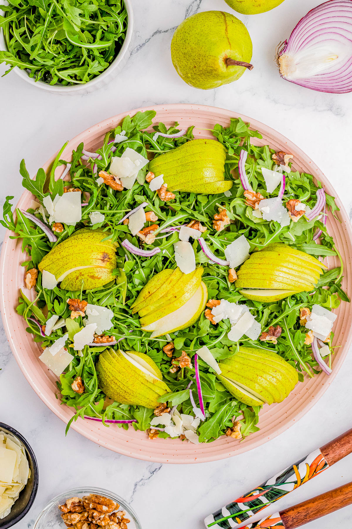 A large salad plate with arugula, sliced pears, red onion, walnuts, and Parmesan cheese. Surroundings include whole pears, a bowl of arugula, a cut red onion, a dish of chopped cheese, and a bowl of walnuts.