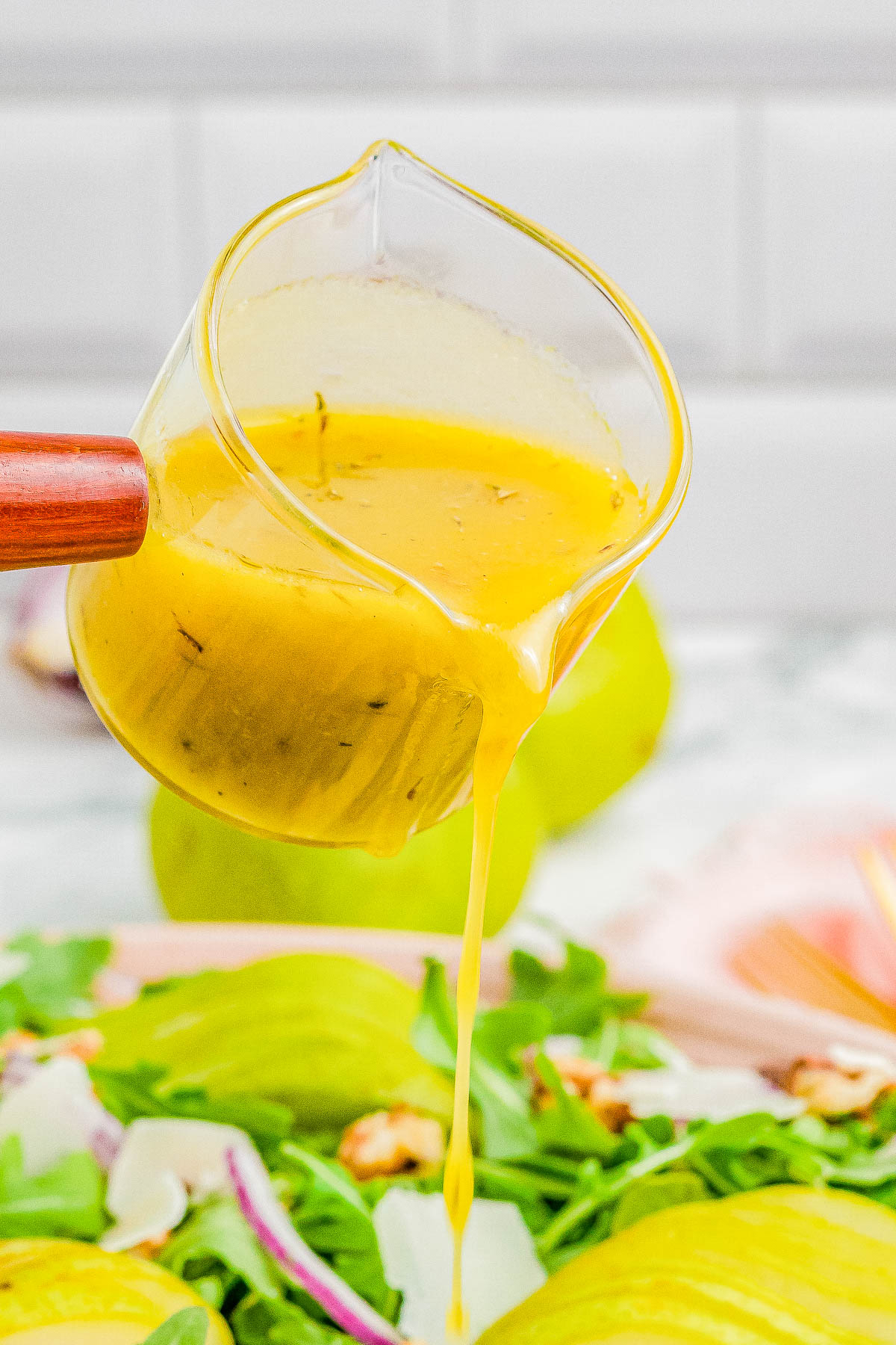 A jug is pouring yellow salad dressing over a bed of arugula, red onion slices, and walnuts.