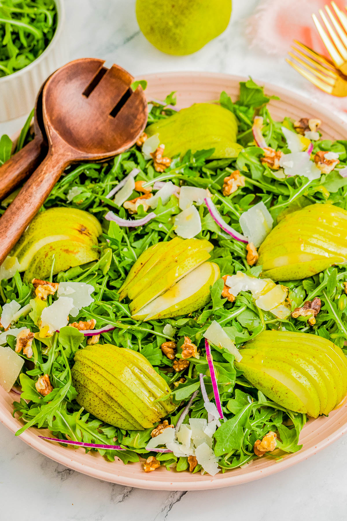 A salad with sliced pears, arugula, walnut pieces, shaved cheese, and red onion slices, served in a round dish with wooden serving utensils.