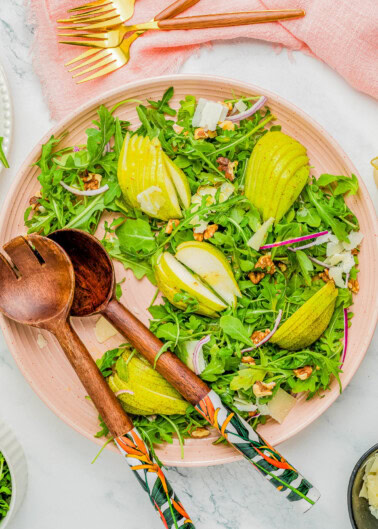 A plate of arugula salad with sliced pears, walnuts, and shaved cheese, accompanied by wooden serving utensils and gold-colored forks on a pink napkin.