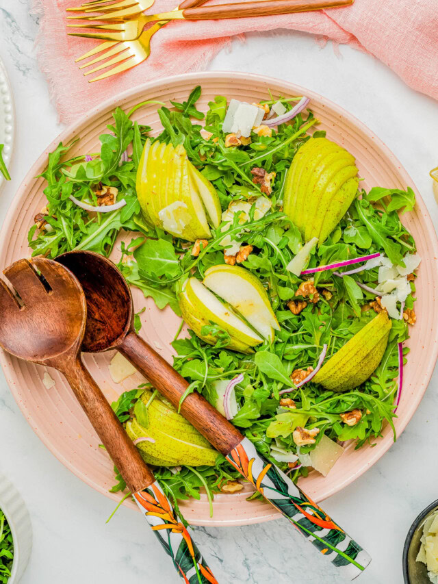 A plate of arugula salad with sliced pears, walnuts, and shaved cheese, accompanied by wooden serving utensils and gold-colored forks on a pink napkin.