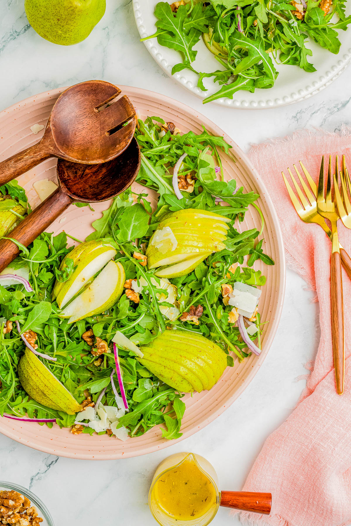 A pear and arugula salad on a pink plate with wooden serving utensils, gold-colored flatware, and a small pitcher of dressing on a pink cloth napkin.