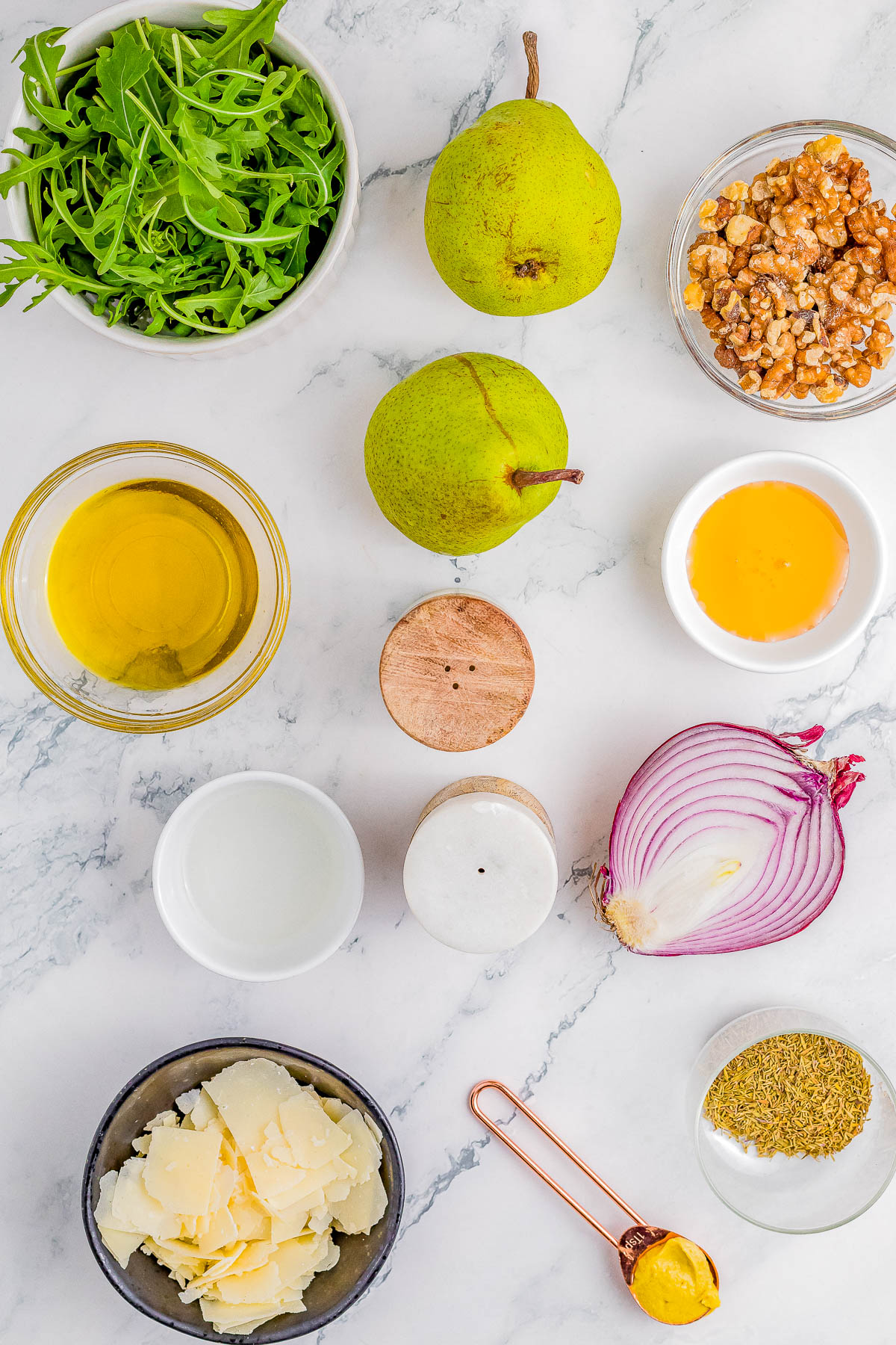 Ingredients for a salad arranged on a marble surface, including arugula, pears, walnuts, olive oil, egg yolk, red onion, cheese, mustard, and seasonings.