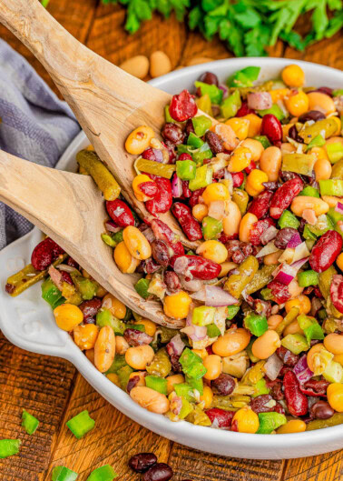 A bowl of mixed bean salad with kidney beans, black beans, corn, green bell peppers, and onions being served with wooden utensils.