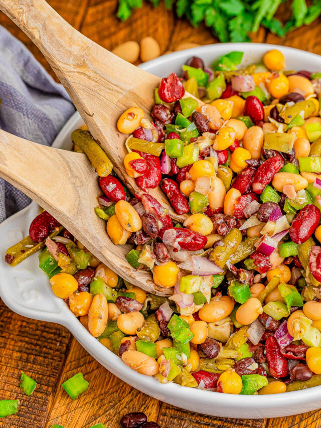 A bowl of mixed bean salad with kidney beans, black beans, corn, green bell peppers, and onions being served with wooden utensils.