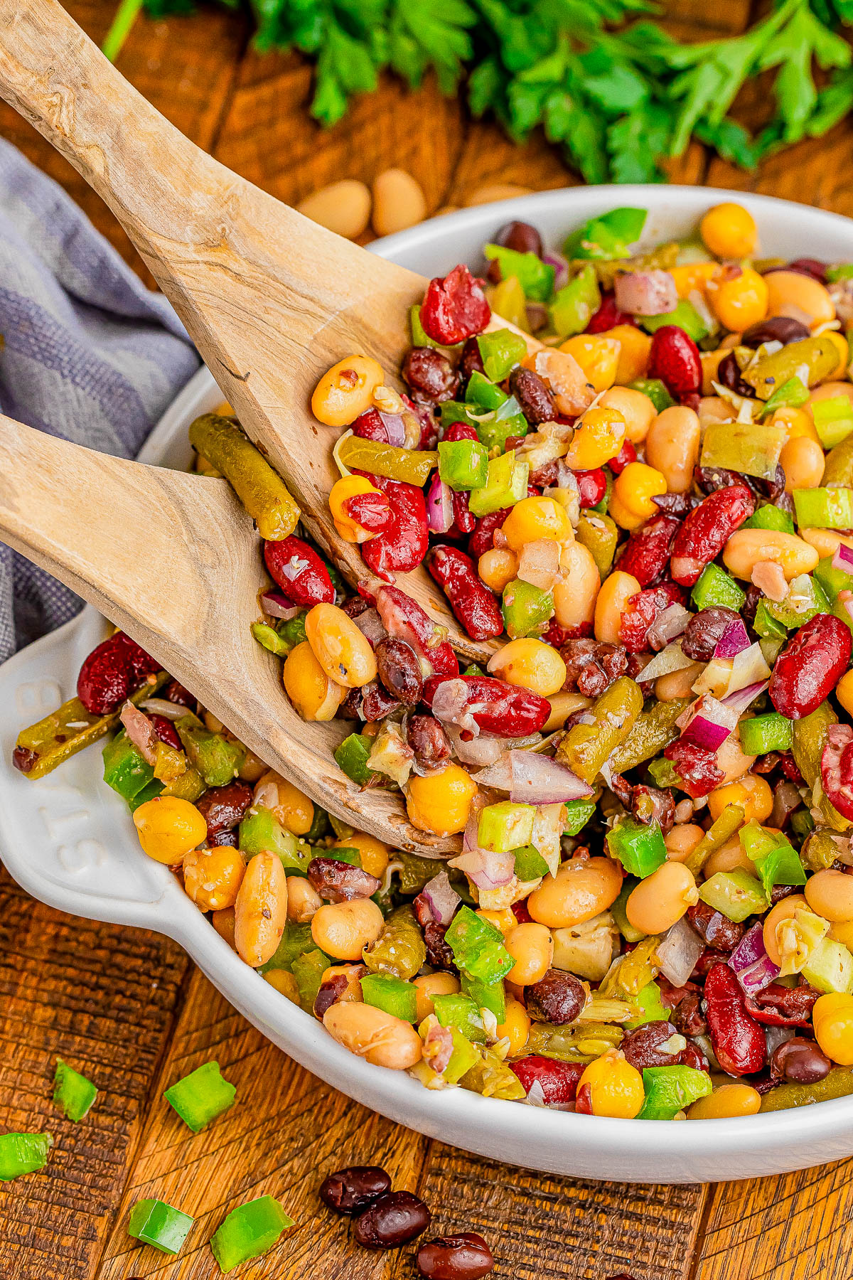 A bowl of mixed bean salad with kidney beans, black beans, corn, green bell peppers, and onions being served with wooden utensils.